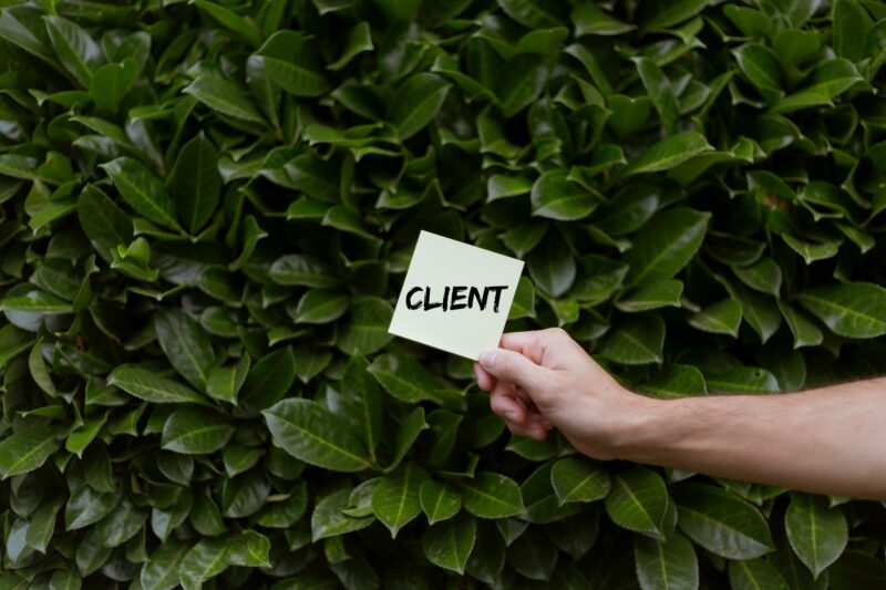 pexels-photo-13092793-13092793 A hand holding a note with the word 'CLIENT' against a lush leafy background.